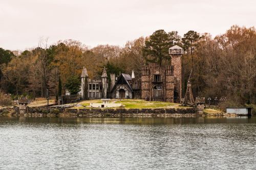 A large, medieval-style castle on a lake, surrounded by trees and a grassy area, under a cloudy sky.