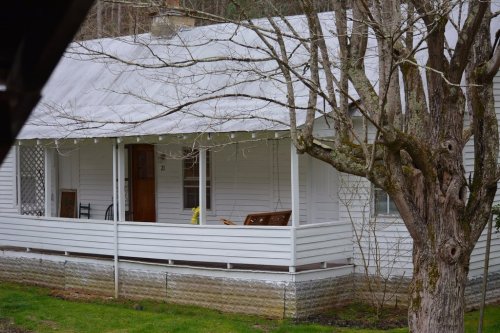 A white house with a porch, surrounded by trees, featuring a bench and a flower pot on the porch.