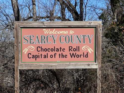Welcome sign for Searcy County, labeled "Chocolate Roll Capital of the World," surrounded by trees.