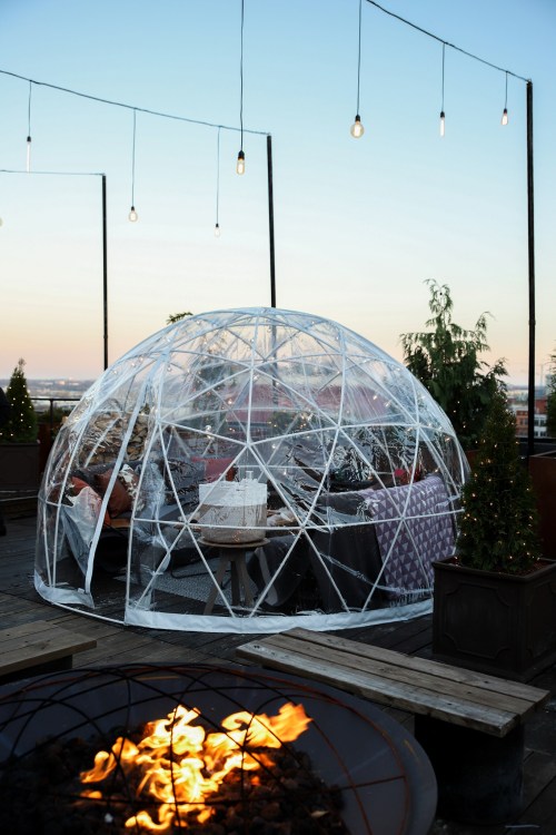 A clear geodesic dome on a rooftop, surrounded by string lights and a fire pit, with a sunset in the background.