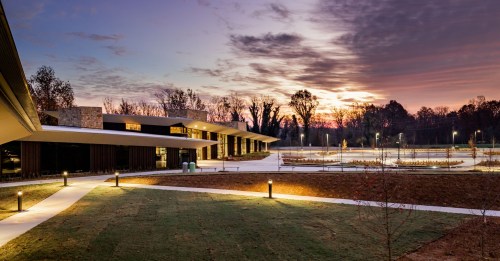 A modern building at sunset, surrounded by landscaped grounds and soft lighting, with trees in the background.