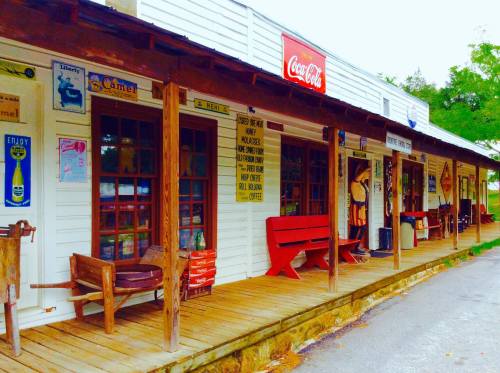 A rustic storefront with wooden benches, vintage signs, and a Coca-Cola advertisement on a sunny day.
