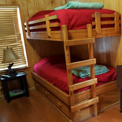 A wooden bunk bed with red bedding, a ladder, and a small nightstand beside it in a cozy room with wooden walls.