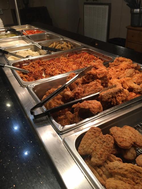 A buffet spread featuring fried chicken, spaghetti, mashed potatoes, and various vegetables in metal trays.