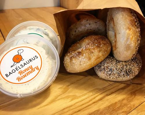 A paper bag filled with assorted bagels next to two containers of honey rosemary cream cheese.