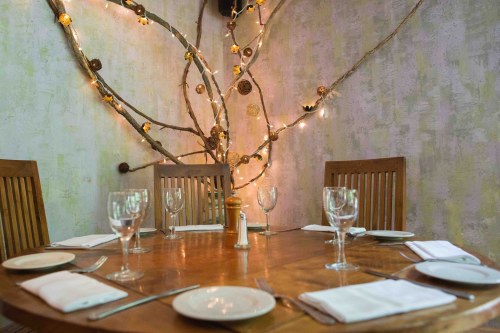 A wooden dining table set for a meal, surrounded by decorative branches and soft lighting on the walls.