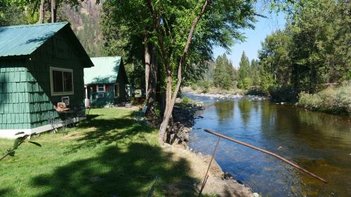 A serene riverside scene featuring green cabins, lush trees, and a calm river under a clear blue sky.