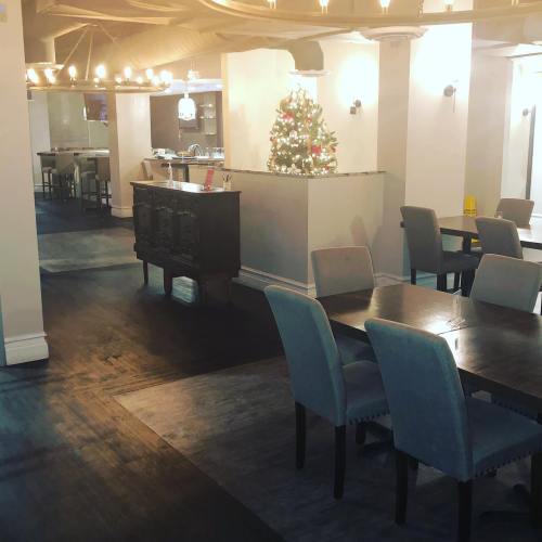 A cozy restaurant interior with tables, a black sideboard, and a small Christmas tree in the corner. Warm lighting.