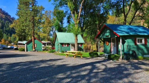 Three green cabins with red accents in a sunny, tree-lined area, surrounded by gravel and a picnic table.