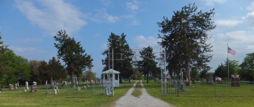 A peaceful cemetery entrance with a gravel path, surrounded by trees and American flags, under a blue sky.