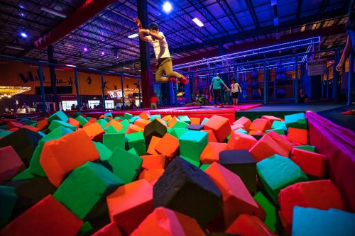 A person jumps into a colorful foam pit at a trampoline park, surrounded by vibrant lighting and other guests.