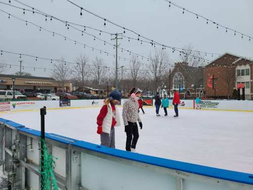 Two skaters on an outdoor ice rink, surrounded by people and festive string lights, with shops in the background.