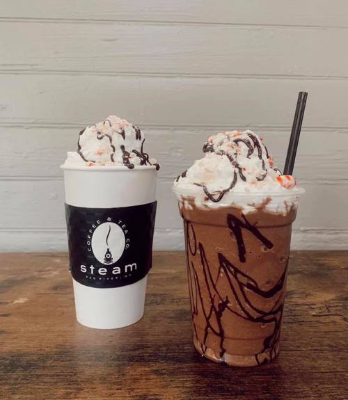 Two coffee drinks on a wooden table: a white cup with a logo and a tall glass topped with whipped cream and chocolate drizzle.