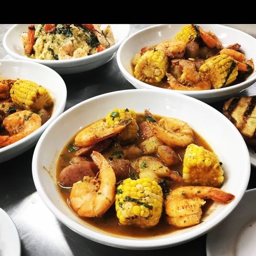 A bowl of shrimp, corn, and sausage in a flavorful broth, surrounded by additional bowls of similar dishes.
