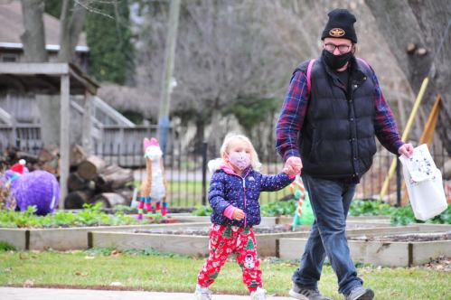 A man and a young girl walk hand in hand outdoors, both wearing masks, surrounded by colorful decorations.