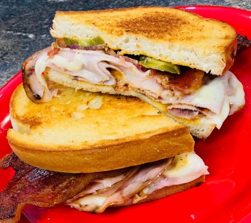 A toasted sandwich with layers of meat, cheese, pickles, and bacon, served on a red plate.