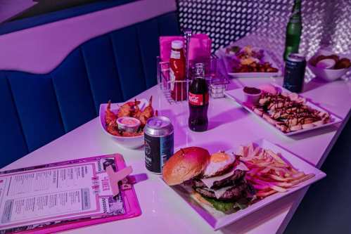 A vibrant table setting with burgers, fries, chicken wings, drinks, and a menu in a colorful diner atmosphere.