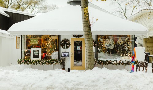 A cozy shop with festive decorations and snow-covered surroundings, featuring a "Merry Christmas" sign and an "Open" sign.
