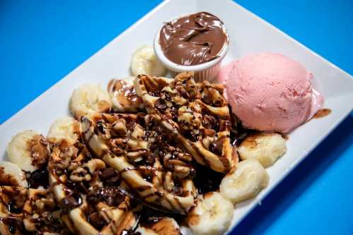 A plate of waffles topped with chocolate sauce, nuts, banana slices, and a scoop of pink ice cream.