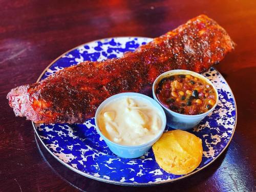 A plate featuring a large, seasoned rib, two side dishes of beans and coleslaw, and a cornbread muffin.