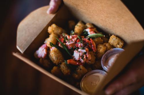 A takeout box filled with lobster, tater tots, and dipping sauces, held by a person.