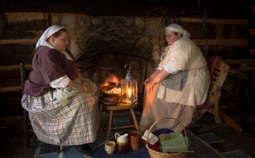 Two women in historical attire sit by a fireplace, preparing food and enjoying a cozy atmosphere in a rustic setting.