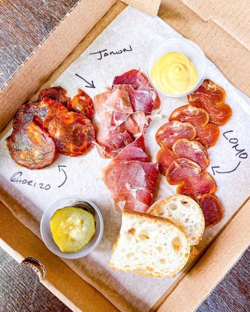 A charcuterie box featuring various meats, pickles, mustard, and slices of bread, labeled with names.