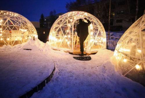 A person enters a lit igloo structure surrounded by snow at dusk, with glowing lights illuminating the scene.