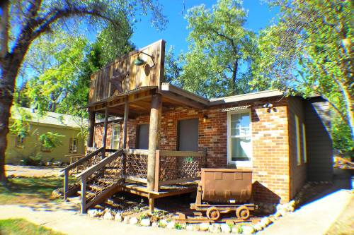 A brick house with a wooden porch, surrounded by trees, featuring a decorative sign and a vintage cart in the yard.