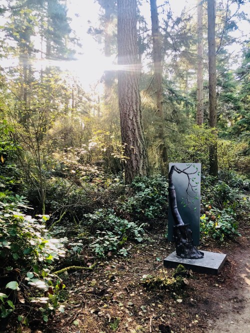 A forest scene with tall trees and sunlight filtering through, featuring a sculpture on a pedestal amidst greenery.