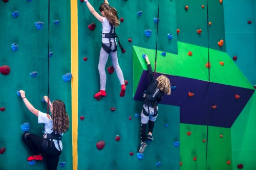 Three children climbing a colorful indoor rock wall, wearing harnesses and focused on reaching the top.
