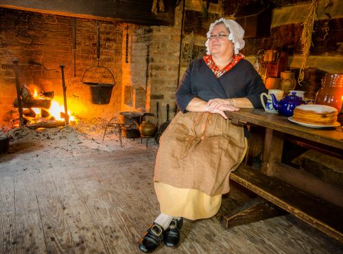 A woman in historical attire sits by a fireplace in a rustic kitchen, with a table featuring baked goods nearby.