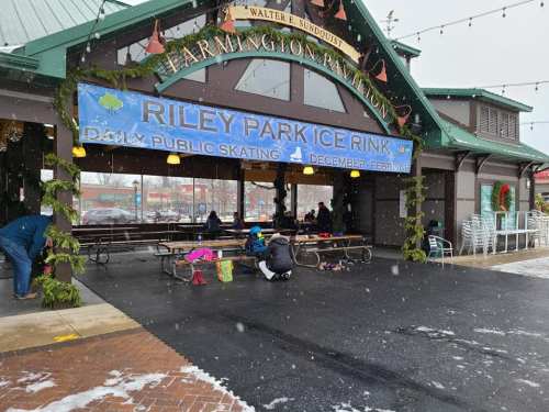 A snowy scene at the Riley Park Ice Rink, featuring a festive banner and people enjoying the outdoor space.