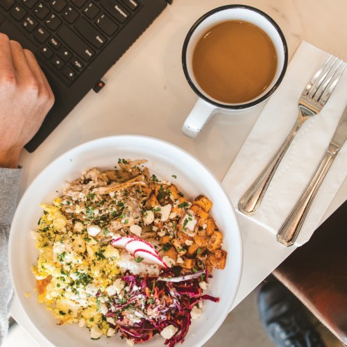 A bowl of colorful food with eggs, chicken, and vegetables, next to a cup of coffee and a keyboard.
