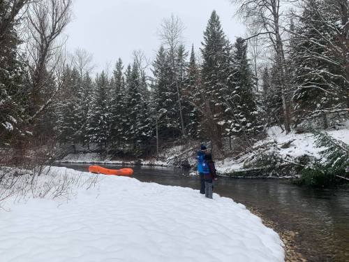 A person stands by a snowy riverbank, with evergreen trees in the background and an orange raft on the water.