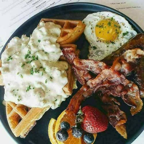 A plate of waffles topped with gravy, a fried egg, crispy bacon, and fresh fruit including strawberries and blueberries.