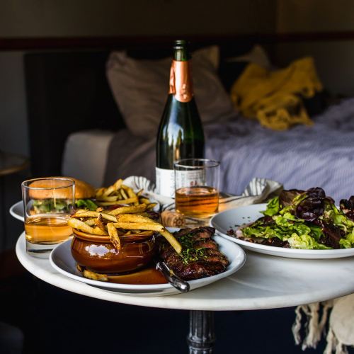 A table with a steak, fries, salad, and drinks, set in a cozy bedroom environment.