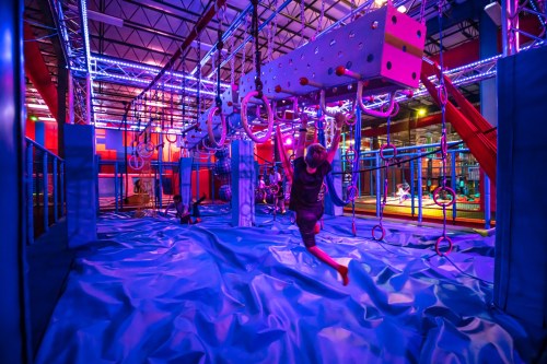 A vibrant indoor play area with colorful lights, featuring climbing structures and soft blue mats. Kids are playing and exploring.