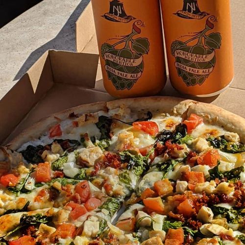 A pizza topped with spinach, tomatoes, and chicken, alongside two cans of IPA beer in orange packaging.