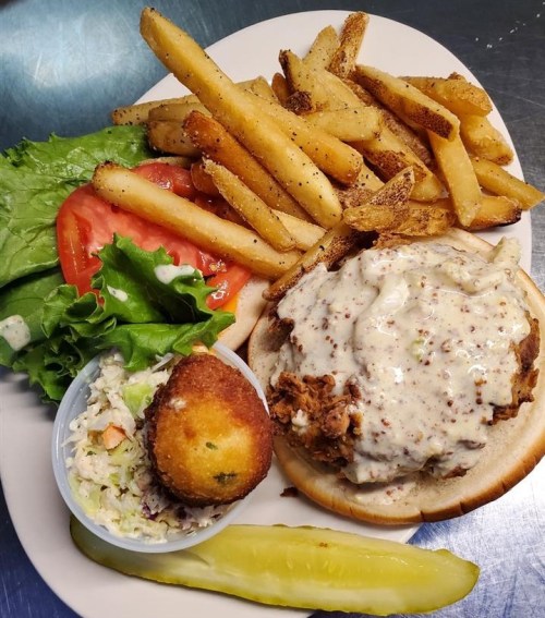 A plate featuring a fried chicken sandwich with creamy sauce, lettuce, tomato, fries, coleslaw, a cornbread muffin, and a pickle.