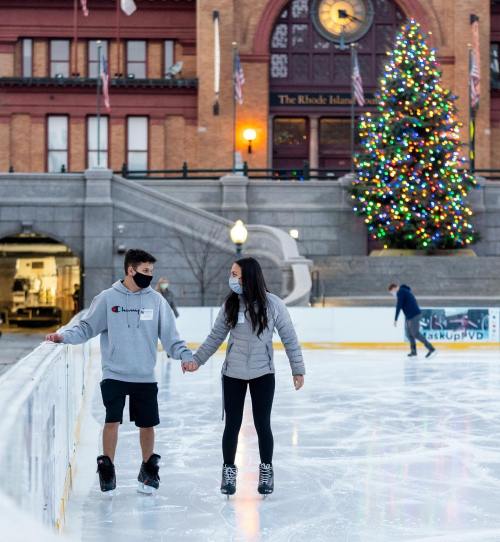 A couple ice skating together on a rink, with a decorated Christmas tree and festive lights in the background.