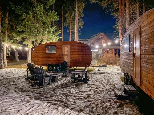A cozy campsite at night featuring wooden trailers, Adirondack chairs, and string lights among tall trees.