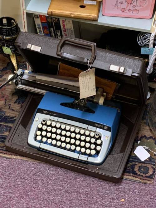 A vintage blue typewriter in a brown case, partially open, with a price tag attached.