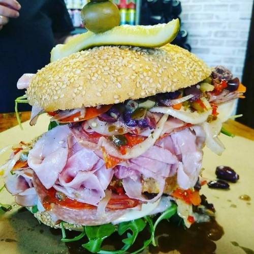 A towering sandwich with layers of meats, olives, and vegetables, topped with a pickle and olive on a sesame seed bun.