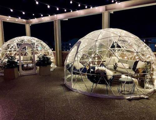 Two illuminated geodesic domes on a rooftop, decorated with cozy seating and string lights at night.