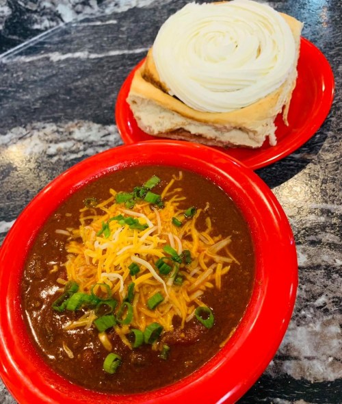 A bowl of chili topped with cheese and green onions, served with a roll of bread on a red plate.