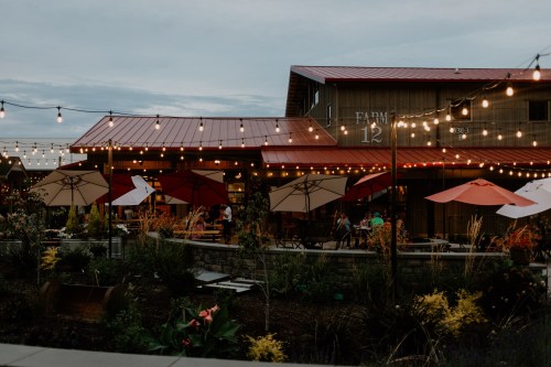 A cozy outdoor dining area with string lights, umbrellas, and plants, set against a rustic building at dusk.