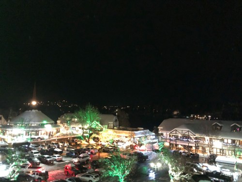 A snowy village at night, illuminated by colorful lights, with cars parked and festive decorations visible.