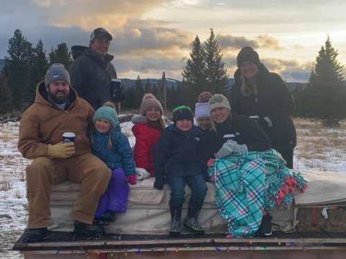 A group of adults and children sitting on a sled in a snowy field, surrounded by trees and mountains in the background.