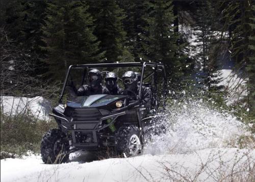 A group of three people in a black off-road vehicle driving through snowy terrain surrounded by trees.